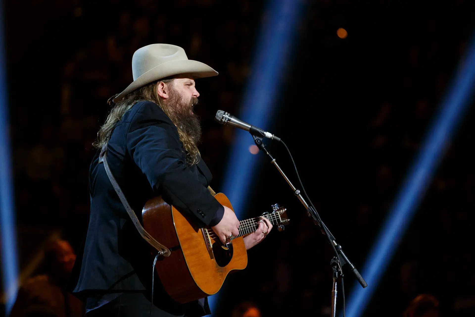 Chris Stapleton singing and playing acoustic guitar at the 2020 Gershwin Prize concert, with blue stage lights behind him.